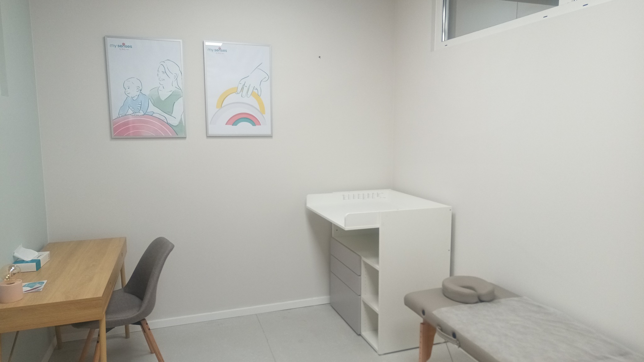 Jasne wnętrze gabinetu z biurkiem, krzesłem, przewijakiem i leżanką do masażu; na ścianie wiszą plakaty z motywami dziecięcymi.