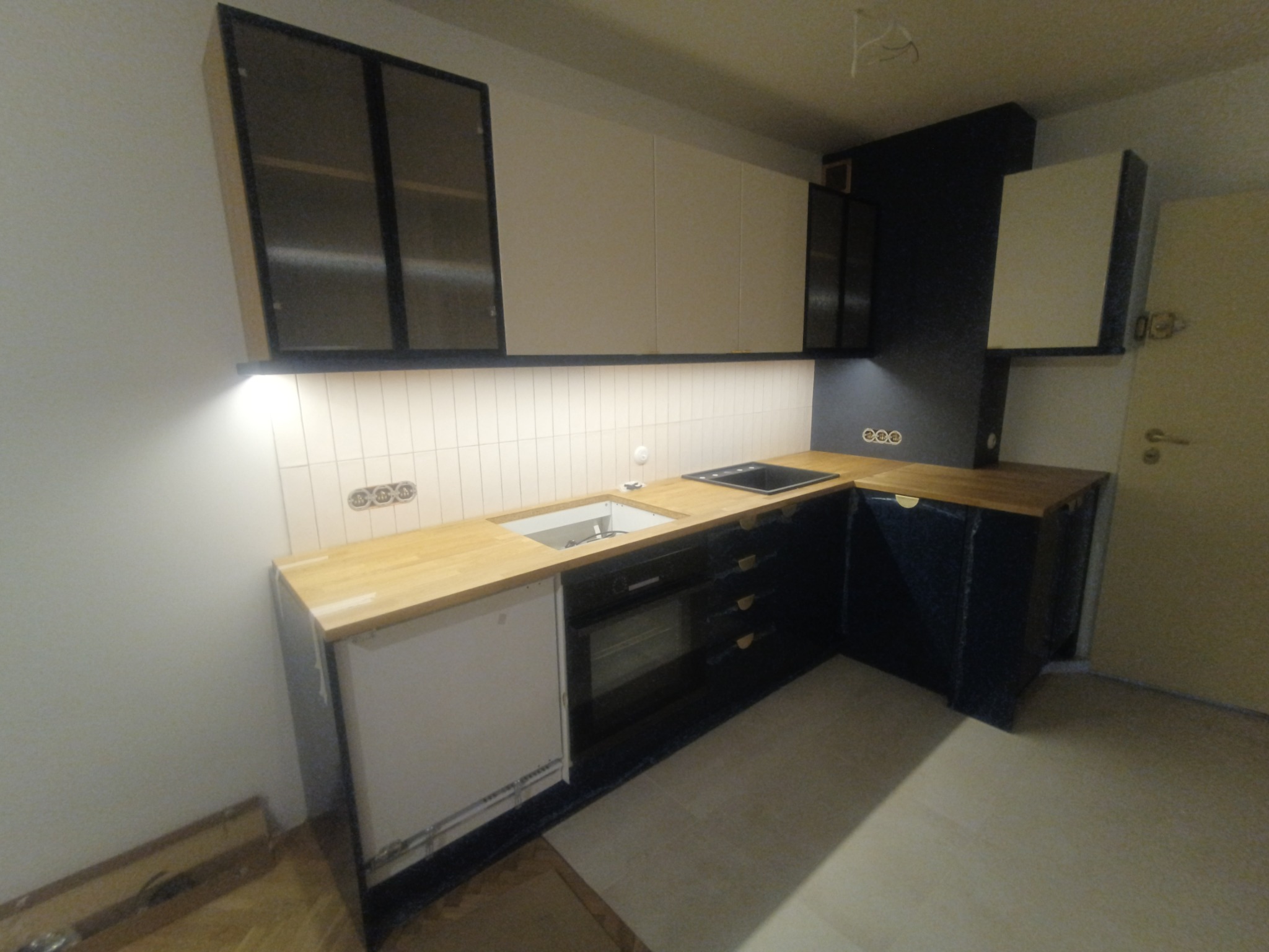 Nowoczesna kuchnia w trakcie montażu: jasne szafki wiszące z czarnymi ramami, drewniany blat i białe płytki na ścianie.