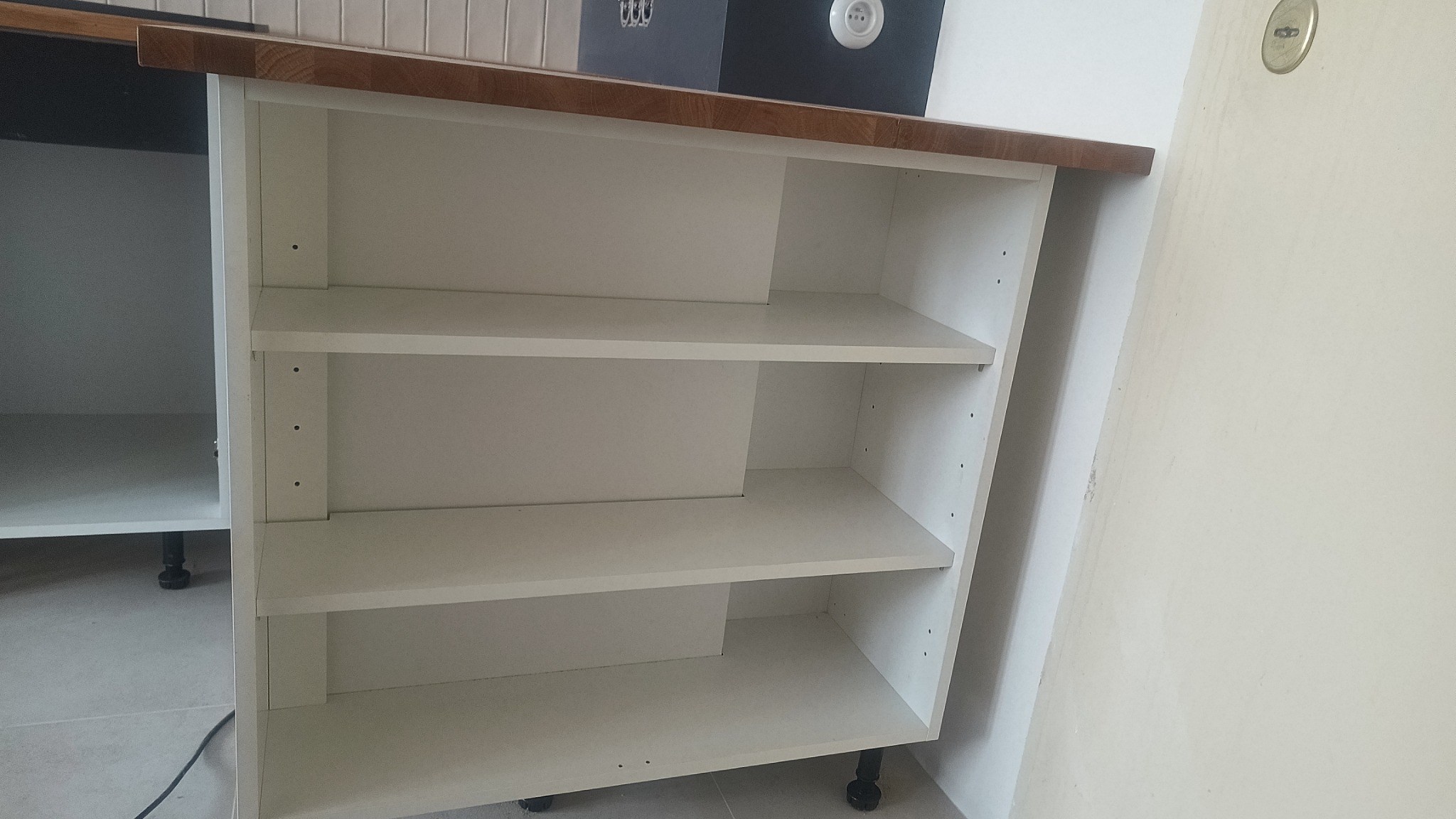 Biała szafka z trzema półkami, zamontowana pod drewnianym blatem kuchennym, widoczna na tle jasnej podłogi i ściany.