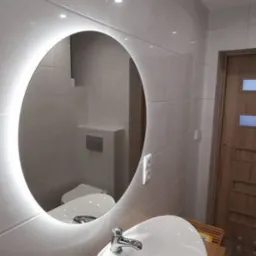 Nowoczesna łazienka z owalnym lustrem podświetlonym LED, białą umywalką nablatową na drewnianym blacie i szarymi płytkami na ścianach.