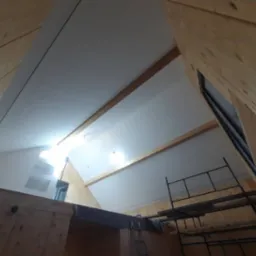 Poddasze z drewnianą konstrukcją i ocieplonym skośnym sufitem, widoczne rusztowanie i okno dachowe w trakcie prac wykończeniowych.