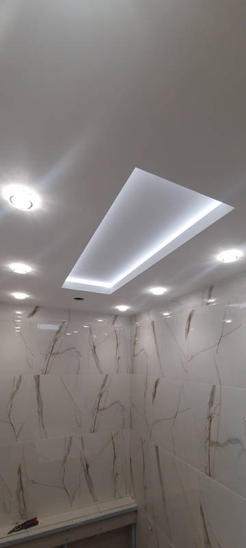 Nowoczesna łazienka z białymi płytkami imitującymi marmur, oświetleniem punktowym i podwieszanym sufitem z oświetleniem LED, na podłodze widoczne narzędzia.