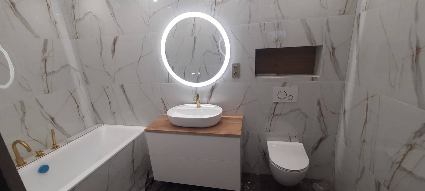 Nowoczesna łazienka z białymi płytkami imitującymi marmur, okrągłym lustrem z podświetleniem LED, białą umywalką na drewnianym blacie, białą wanną z złotą armaturą i podwieszaną toaletą.