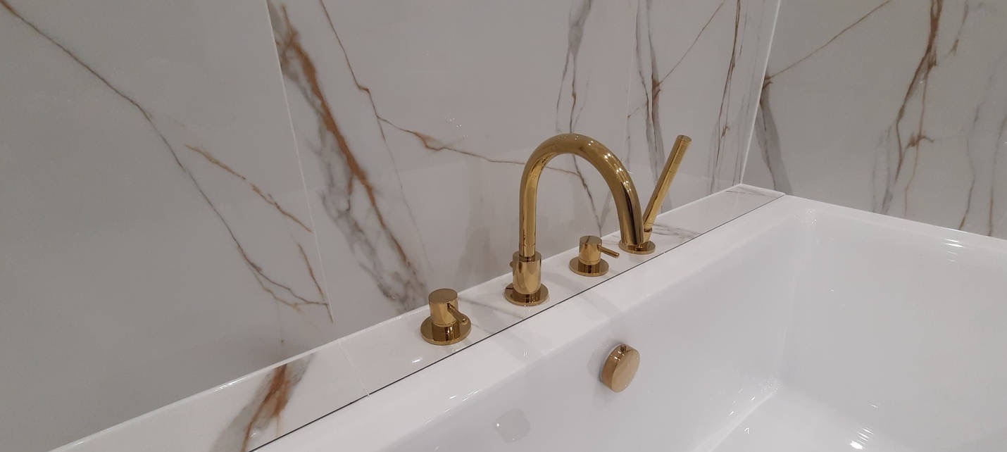Bateria wannowa w kolorze złotym, zamontowana na wannie z białego akrylu, tło stanowi ściana wyłożona płytkami imitującymi marmur.
