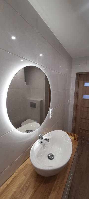 Nowoczesna łazienka z owalnym lustrem podświetlonym LED, białą umywalką nablatową na drewnianym blacie i szarymi płytkami na ścianach.