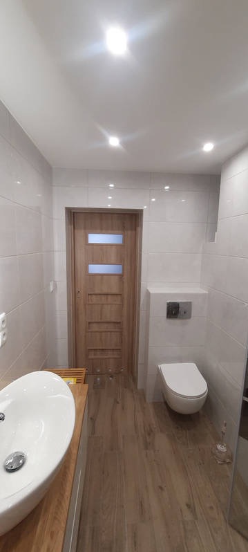 Nowoczesna łazienka z białymi płytkami ściennymi, drewnopodobną podłogą, wiszącą toaletą z chromowanym przyciskiem i umywalką nablatową na drewnianym blacie, drzwi z przeszkleniami w tle.