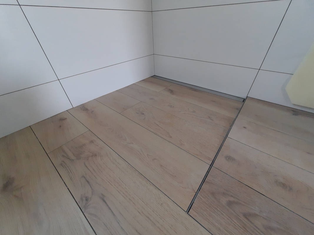 Jasne panele podłogowe ułożone w rogu pomieszczenia z białymi, kwadratowymi płytkami na ścianach i widocznym odpływem liniowym.