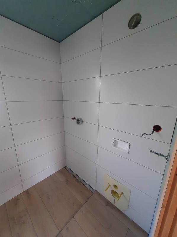 Wnętrze łazienki w trakcie wykańczania: ułożone białe, poziome płytki, drewnopodobna podłoga, widoczne wyprowadzenia instalacji wodnej i elektrycznej oraz stelaż podtynkowy WC.