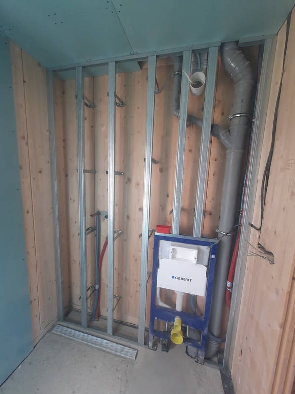 Wnętrze łazienki w trakcie remontu z widoczną konstrukcją metalową pod zabudowę, drewnianą boazerią, częściowo położonymi płytami gipsowo-kartonowymi i zamontowanym stelażem podtynkowym Geberit...