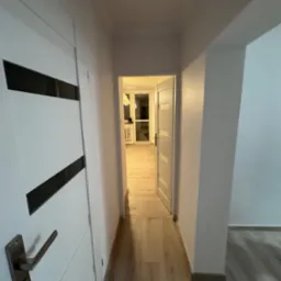 Wnętrze mieszkania po remoncie: widok na przedpokój z białymi drzwiami z czarnymi wstawkami, jasną podłogą i widokiem na pokój w tle. Drzwi po prawej stronie są otwarte.