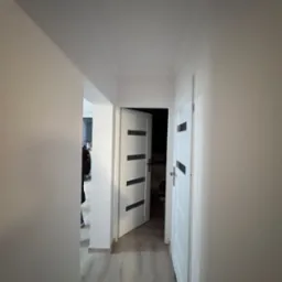 Korytarz po remoncie z pomalowanymi ścianami na jasny kolor, widoczne dwoje białych drzwi z czarnymi paskami i fragment podłogi z paneli.