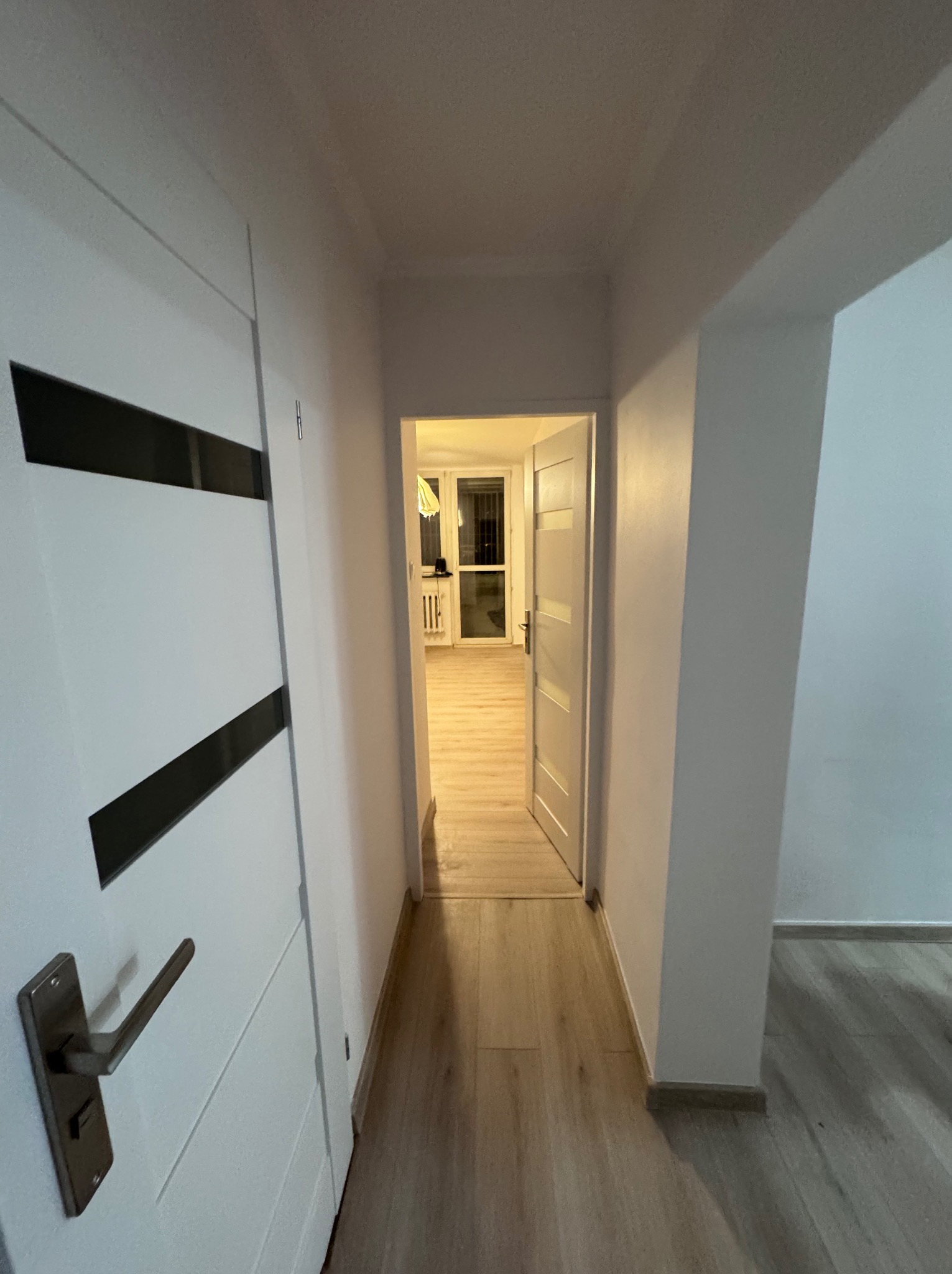 Wnętrze mieszkania po remoncie: widok na przedpokój z białymi drzwiami z czarnymi wstawkami, jasną podłogą i widokiem na pokój w tle. Drzwi po prawej stronie są otwarte.