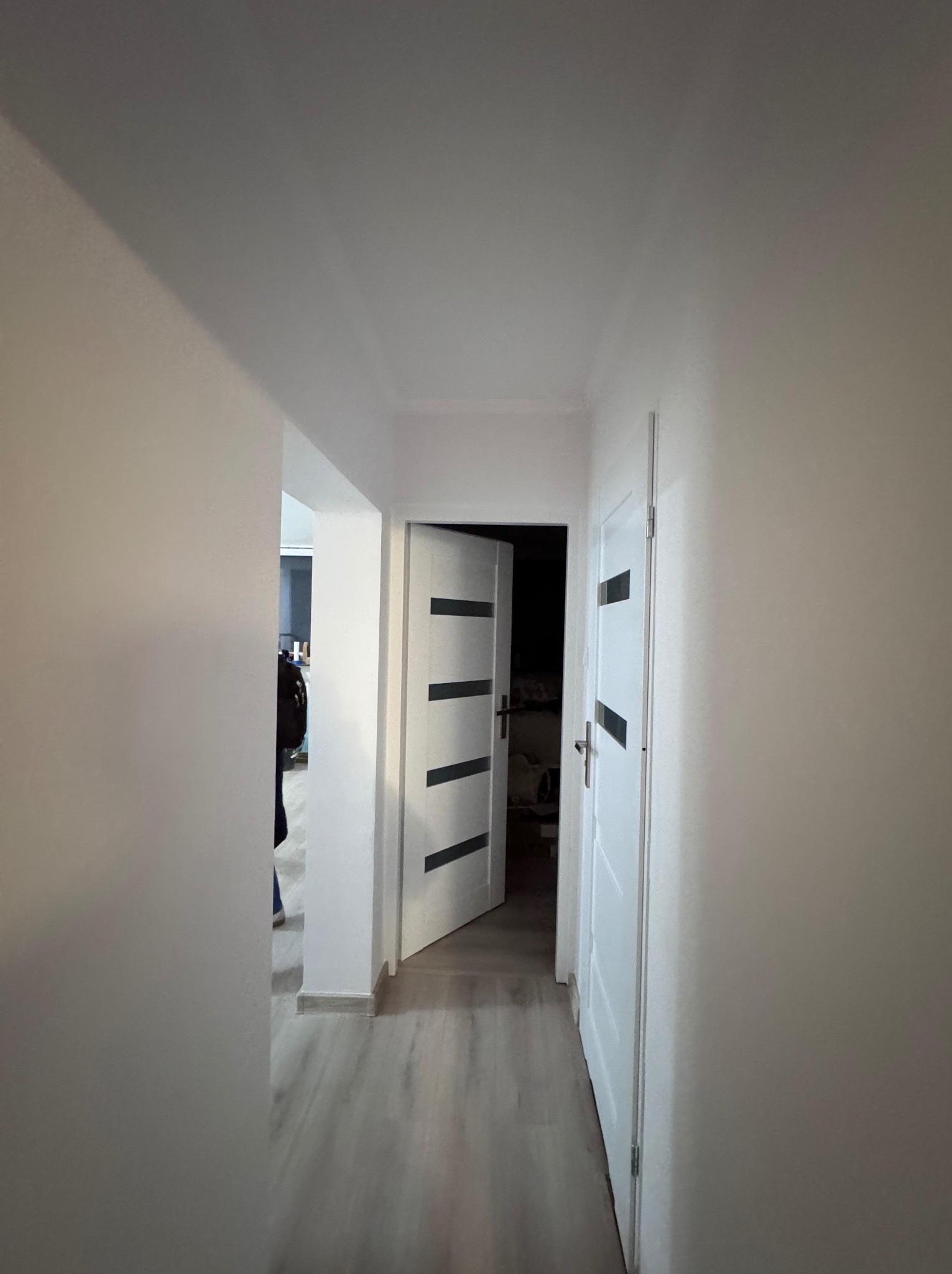 Korytarz po remoncie z pomalowanymi ścianami na jasny kolor, widoczne dwoje białych drzwi z czarnymi paskami i fragment podłogi z paneli.
