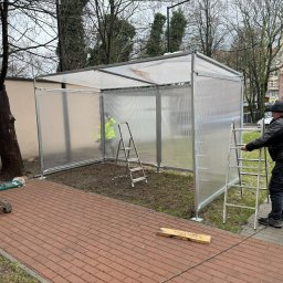 OMM 4x4 Robert Boruszak - Montaż zadaszenia z przezroczystych paneli na metalowej konstrukcji. Pracownik na drabinie, narzędzia na palecie obok, widoczne budynki w tle.