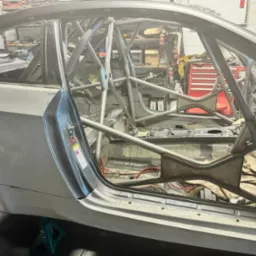 Wnętrze samochodu wyścigowego w trakcie modyfikacji, z widoczną klatką bezpieczeństwa z rur stalowych, spawane elementy konstrukcyjne, częściowo zdemontowane wnętrze, okablowanie i narzędzia w tle.