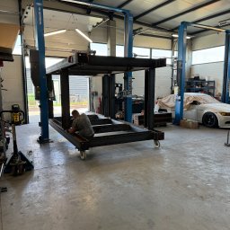 OMM 4x4 Robert Boruszak - Warsztat z surową, metalową konstrukcją na kółkach w trakcie tworzenia, spawacz w koszulce moro pracuje nad spoinami, w tle podnośniki samochodowe i częściowo zakryty samochód.