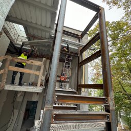 OMM 4x4 Robert Boruszak - Montaż stalowej konstrukcji schodów zewnętrznych na placu budowy, widoczni pracownicy w odblaskowych kamizelkach. Surowy stan budynku w tle.