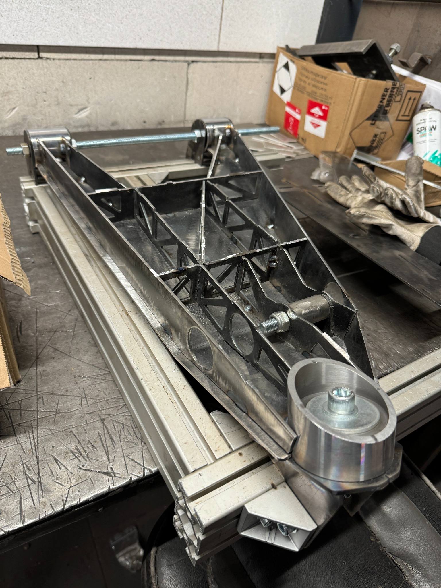 Metalowa konstrukcja z elementami toczonymi i gwintowanymi, prawdopodobnie fragment większego mechanizmu, widoczna na stole warsztatowym z narzędziami i akcesoriami.