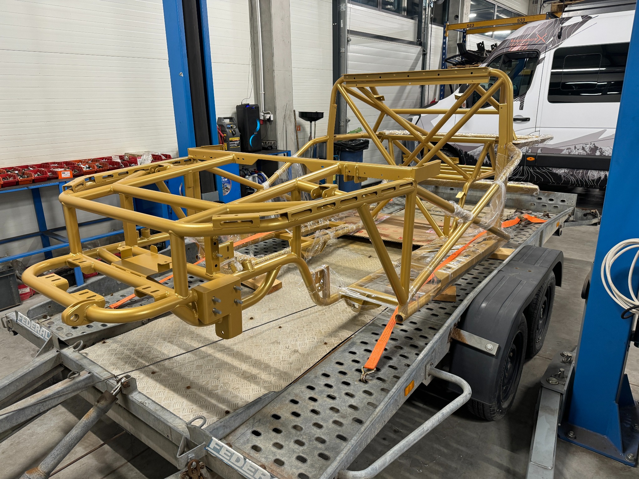 Złoty, rurowy szkielet konstrukcji pojazdu, zabezpieczony folią, umieszczony na metalowej przyczepie transportowej w warsztacie.