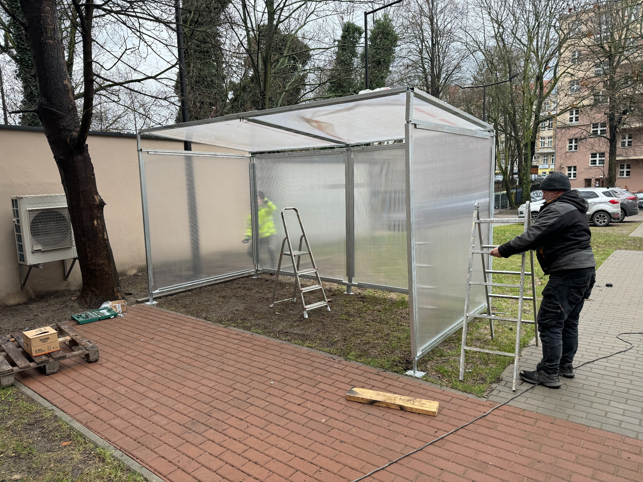 Montaż zadaszenia z przezroczystych paneli na metalowej konstrukcji. Pracownik na drabinie, narzędzia na palecie obok, widoczne budynki w tle.