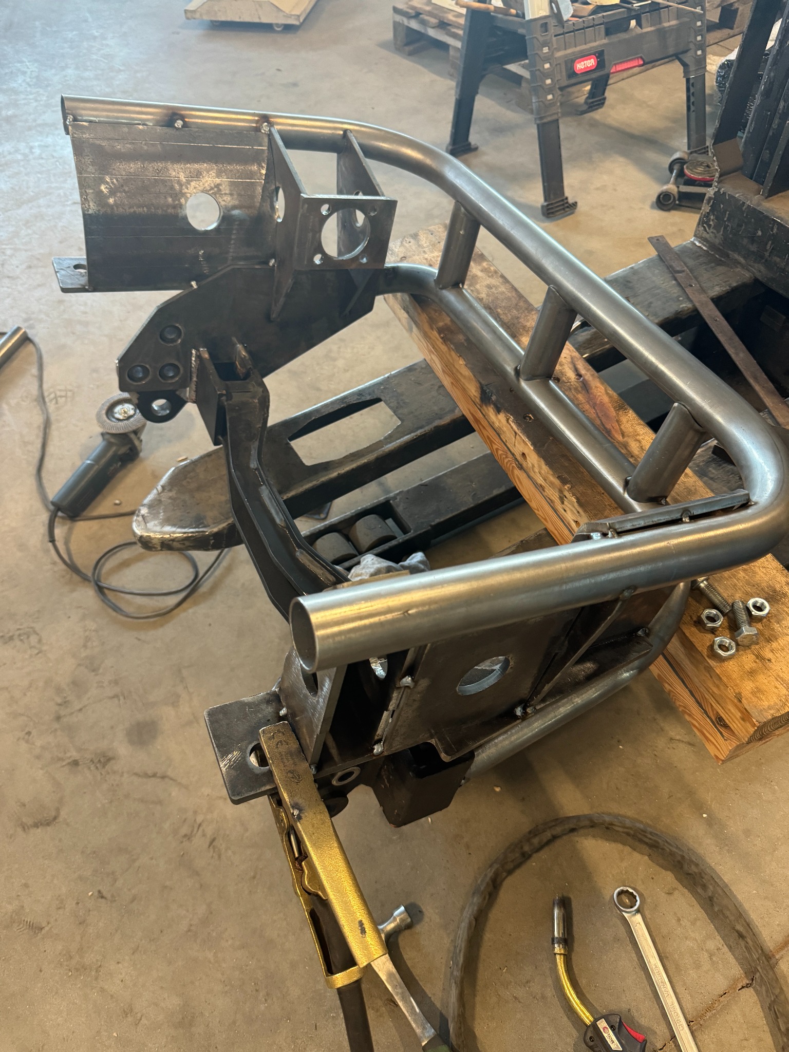 Metalowa konstrukcja z rur i blach w trakcie spawania, widoczne narzędzia ślusarskie, w tym spawarka i zaciski, na tle betonowej podłogi warsztatu.