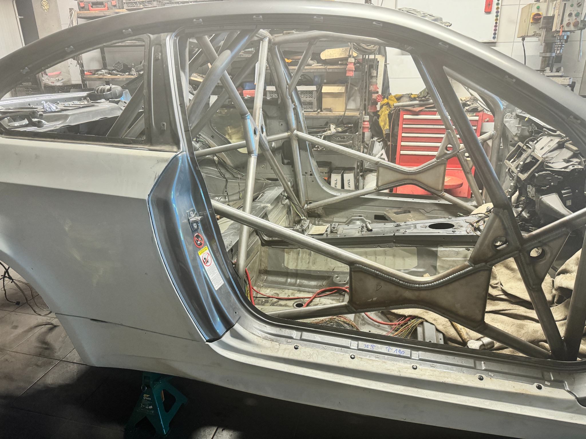 Wnętrze samochodu wyścigowego w trakcie modyfikacji, z widoczną klatką bezpieczeństwa z rur stalowych, spawane elementy konstrukcyjne, częściowo zdemontowane wnętrze, okablowanie i narzędzia w tle.