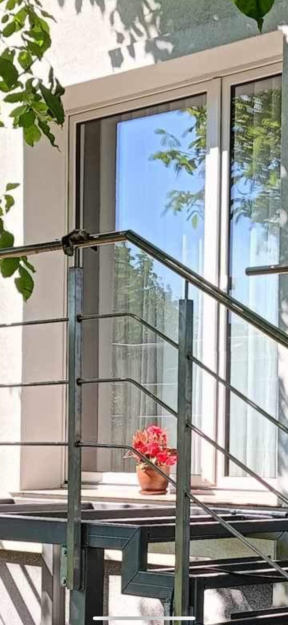 Zewnętrzne schody z metalową konstrukcją i balustradą, z doniczką z czerwonymi kwiatami na podeście, widok na okno i zieleń w tle.
