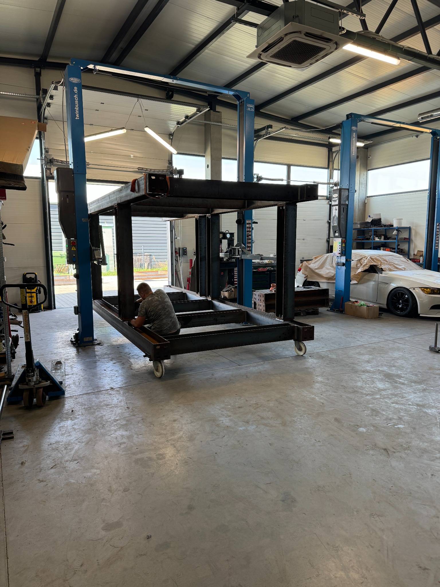 Warsztat z surową, metalową konstrukcją na kółkach w trakcie tworzenia, spawacz w koszulce moro pracuje nad spoinami, w tle podnośniki samochodowe i częściowo zakryty samochód.