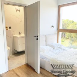 Yury Hancharyk - Widok na sypialnię z łazienką en-suite, z otwartymi drzwiami ukazującymi umywalkę na szafce i toaletę, drewniana podłoga, duże okno z widokiem na zieleń, minimalistyczny design.