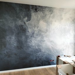 Yury Hancharyk - Ściana w odcieniach szarości z efektem dekoracyjnym przypominającym chmury, widoczne gniazdka elektryczne i kartony z elementami wyposażenia, podłoga drewnopodobna.