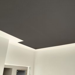 Yury Hancharyk - Pomieszczenie po remoncie, sufit w kolorze grafitowym z wbudowanym oświetleniem LED, białe ściany, widoczne narzędzia i drzwi w tle.