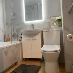 Nowoczesna łazienka z marmurowymi płytkami, podświetlanym lustrem nad białą szafką umywalkową, toaletą typu compact i szarym dywanikiem na drewnopodobnej podłodze.