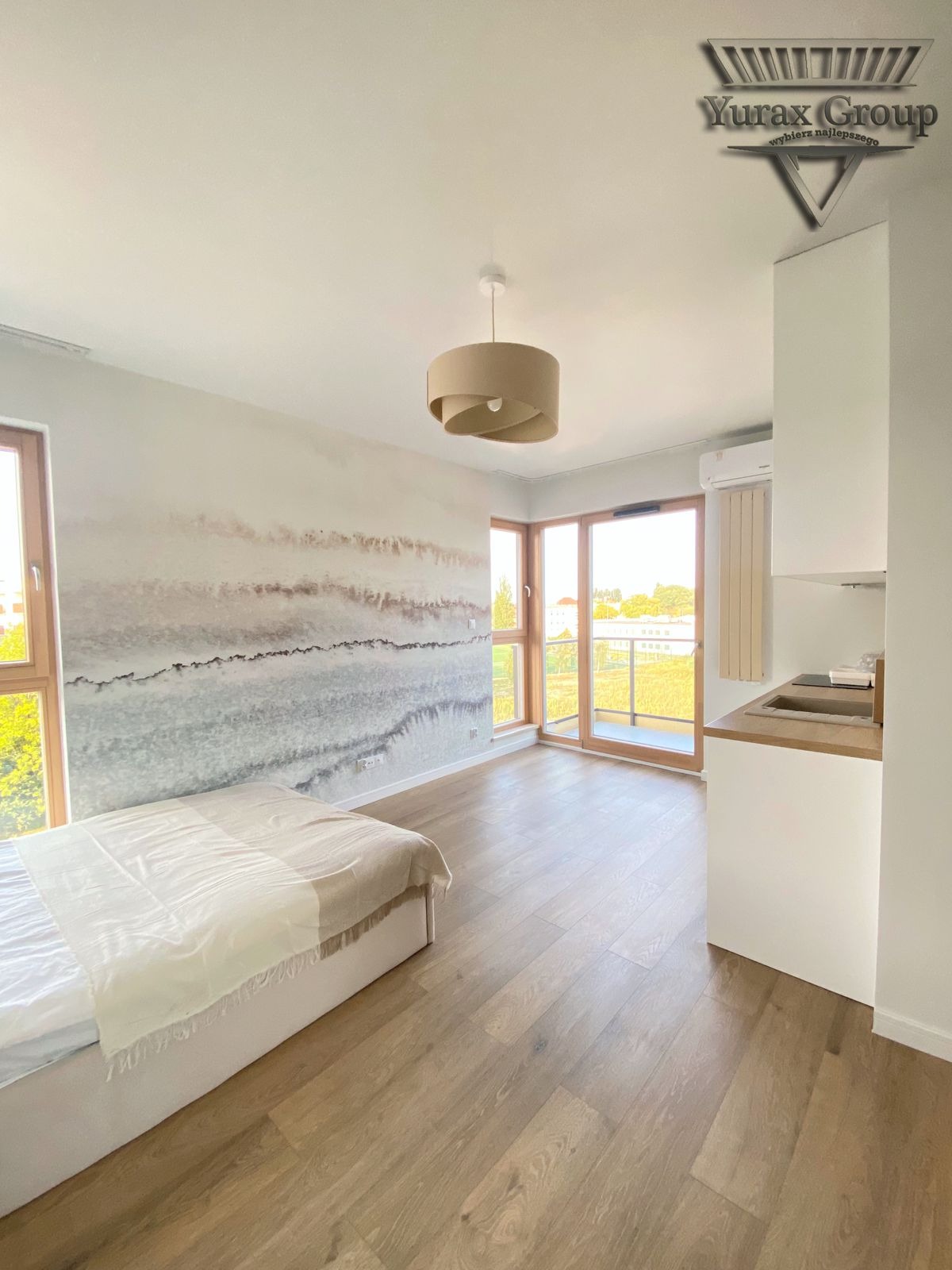 Jasne wnętrze pokoju z łóżkiem, fototapetą w odcieniach szarości i beżu na ścianie, drewnianą podłogą, oknami balkonowymi i lampą w stylu skandynawskim; widoczny fragment aneksu kuchennego...