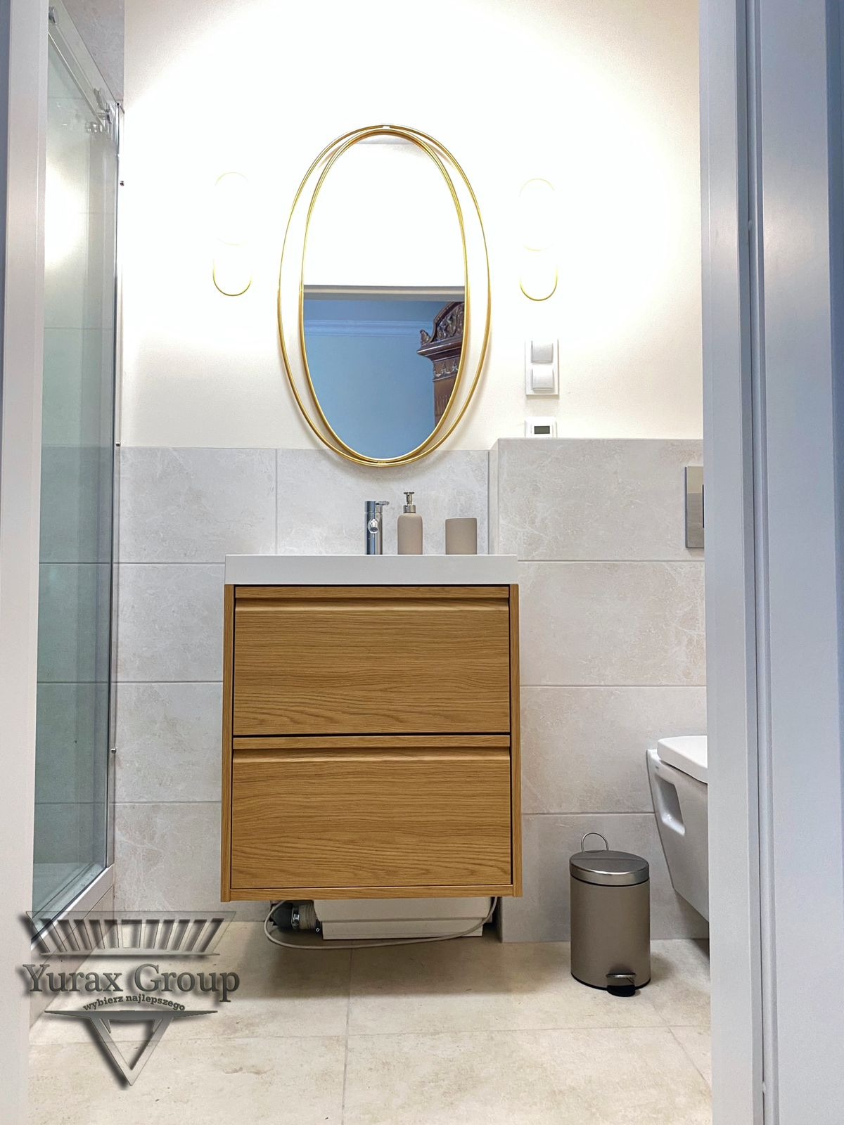 Nowoczesna łazienka z wiszącą szafką z drewna, owalne lustro w złotej ramie, szare płytki i widoczny fragment kabiny prysznicowej.