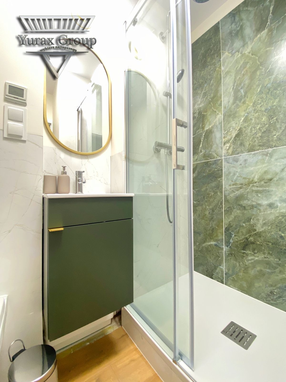 Nowoczesna łazienka z zielonymi akcentami: kabina prysznicowa z marmurowymi płytkami, umywalka z szafką w odcieniu butelkowej zieleni i owalne lustro w złotej ramie, uzupełnione o logo 'Yurax Group'.