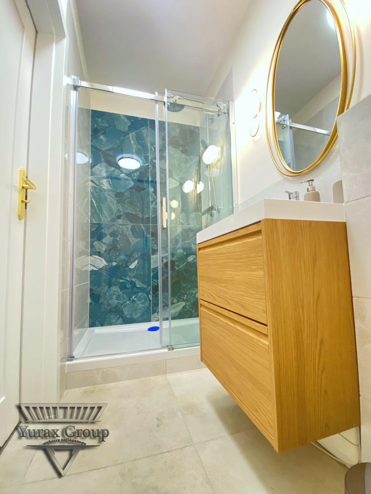 Nowoczesna łazienka z kabiną prysznicową z zielonymi marmurowymi płytkami, umywalką z drewnianą szafką i owalnym lustrem w złotej ramie. Logo firmy widoczne na podłodze.