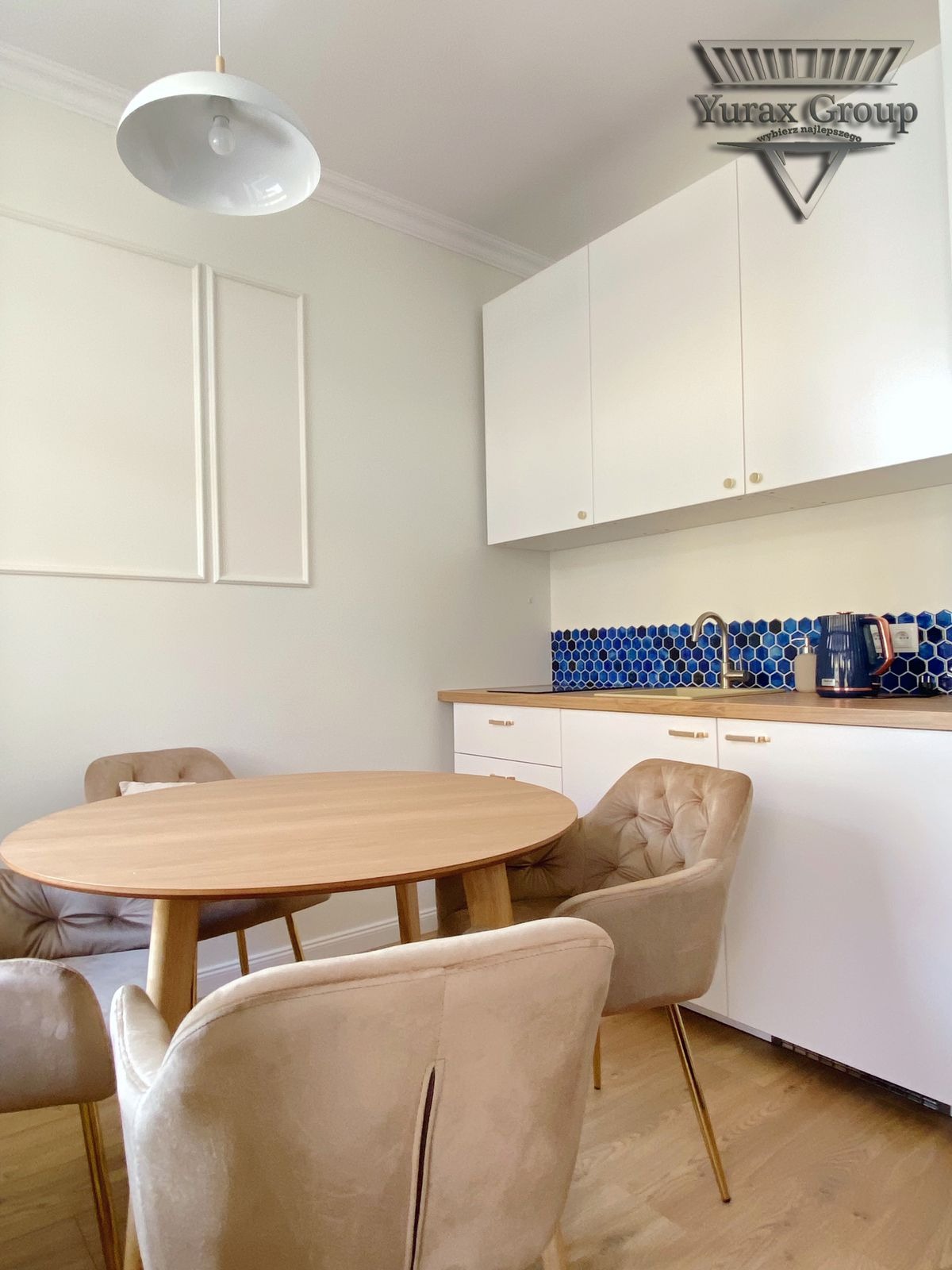 Jasna kuchnia z okrągłym drewnianym stołem i beżowymi krzesłami z pikowaniem, niebieska mozaika nad blatem, białe szafki z złotymi uchwytami, logo firmy na górnej szafce.