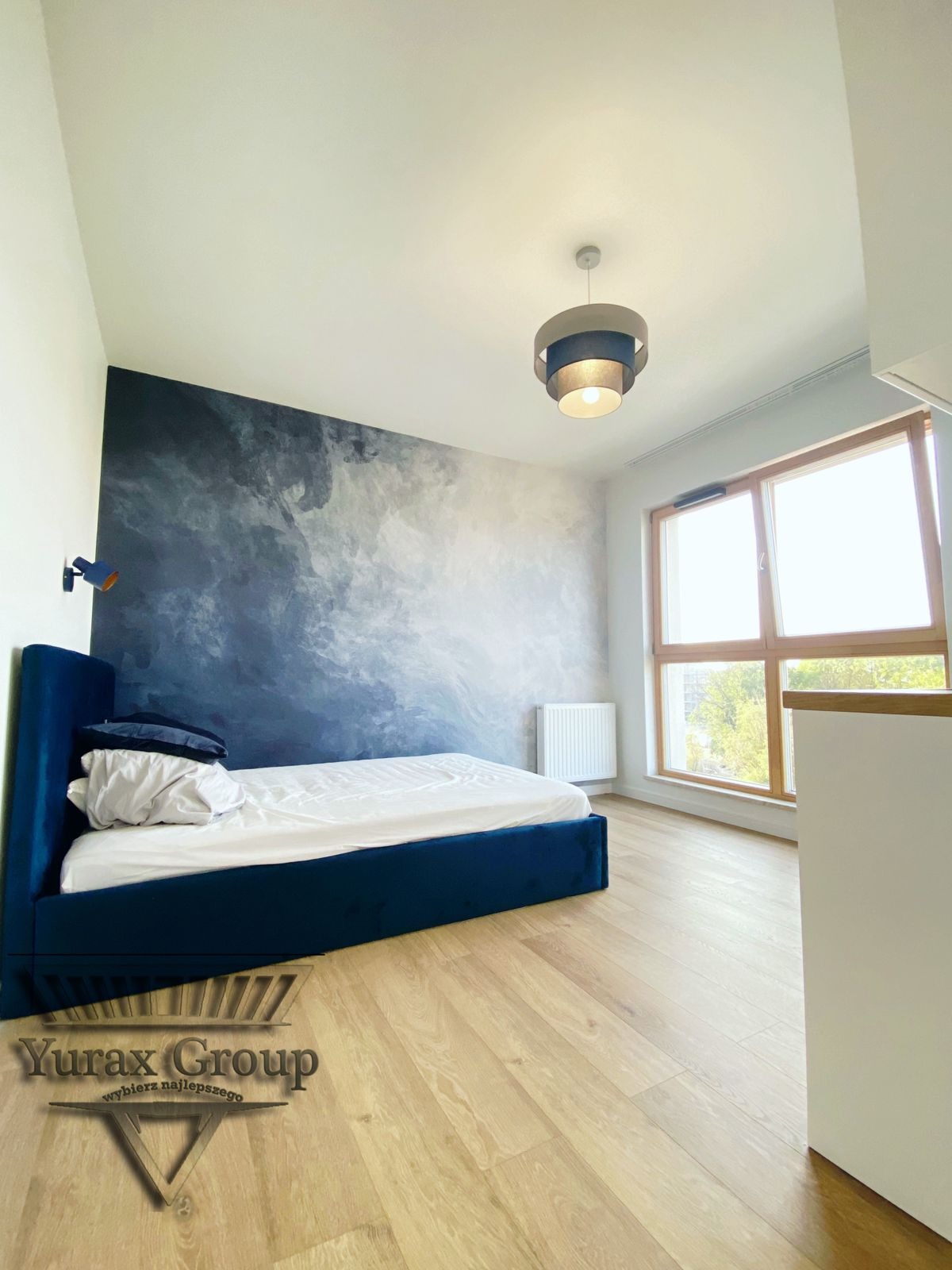 Sypialnia z łóżkiem w obiciu z granatowego weluru, ściana z efektem malarskim w odcieniach błękitu i szarości, drewniana podłoga i lampa sufitowa z trzema pierścieniami.