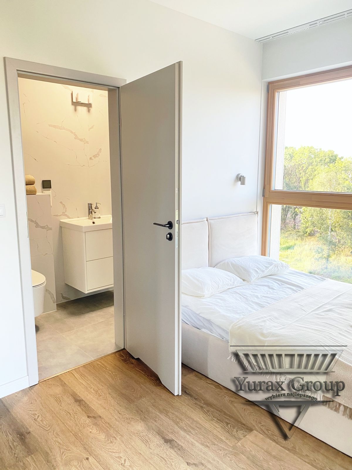 Widok na sypialnię z łazienką en-suite, z otwartymi drzwiami ukazującymi umywalkę na szafce i toaletę, drewniana podłoga, duże okno z widokiem na zieleń, minimalistyczny design.