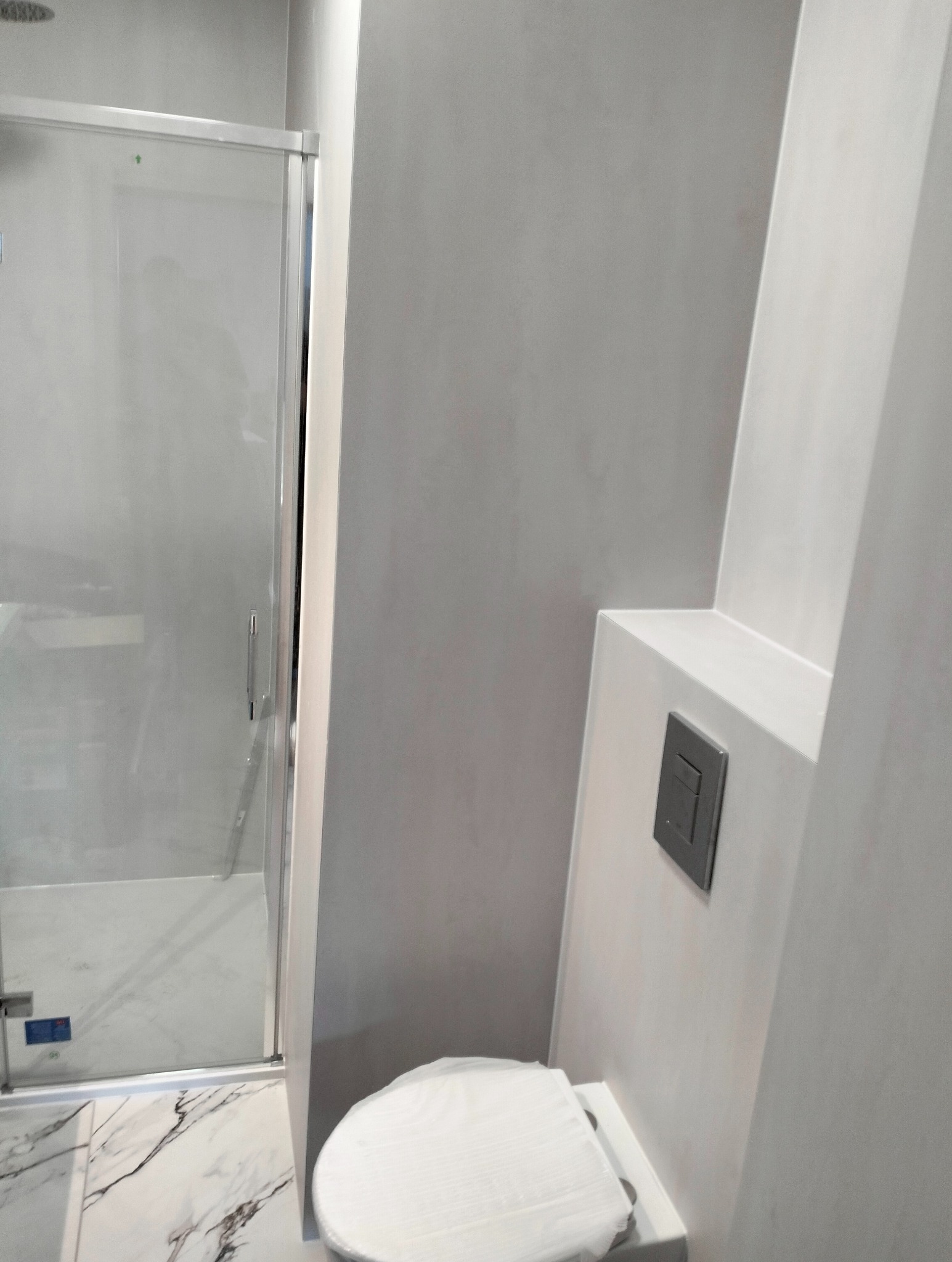 Nowoczesna łazienka w odcieniach szarości i bieli z kabiną prysznicową ze szklanymi drzwiami oraz podwieszaną toaletą ze spłuczką w kolorze grafitowym, wykończona marmurową posadzką.