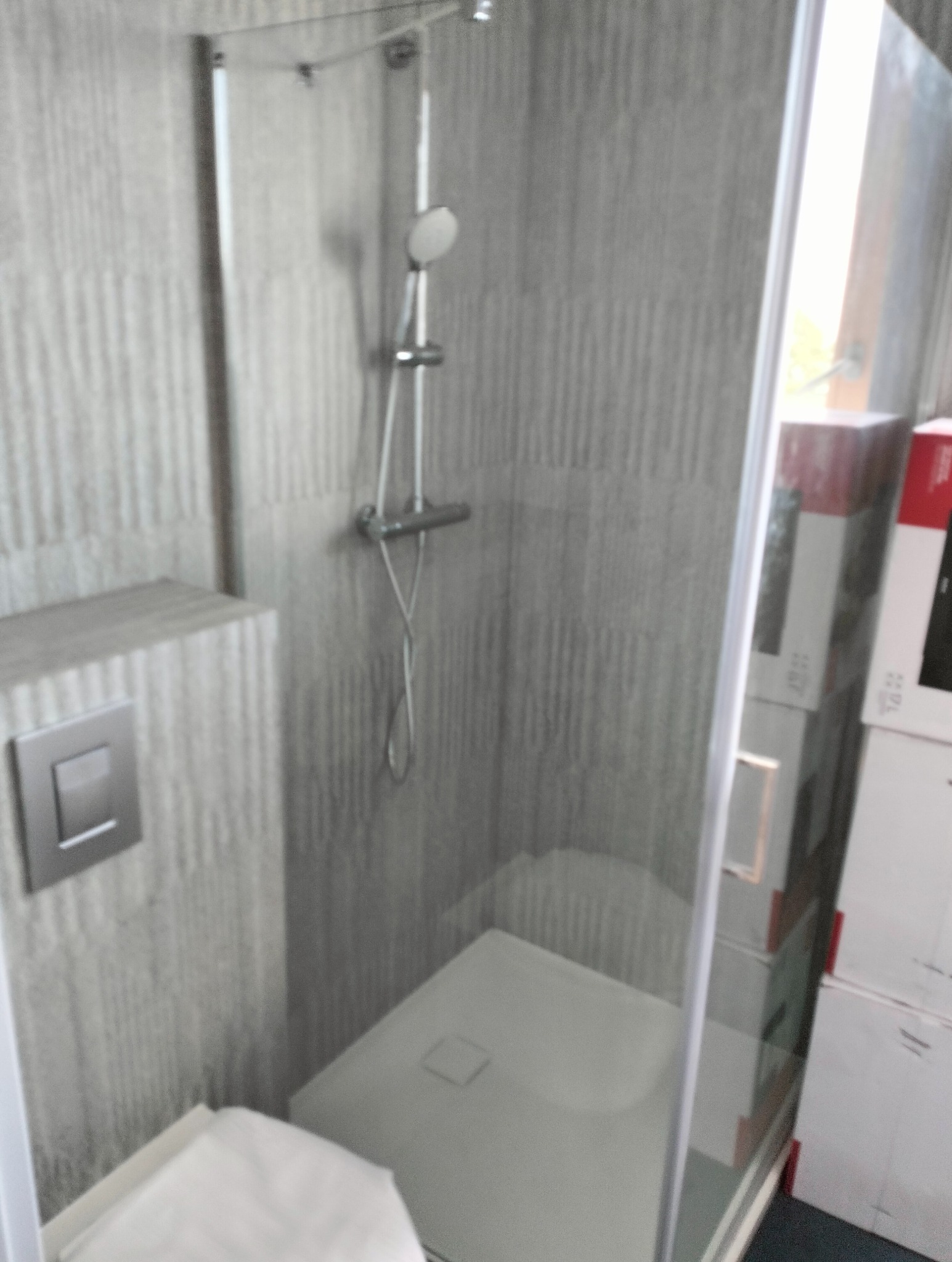 Nowoczesna kabina prysznicowa z deszczownicą, płytki imitujące pionowe pasy, szklana ściana kabiny, widoczne pudełka w tle.