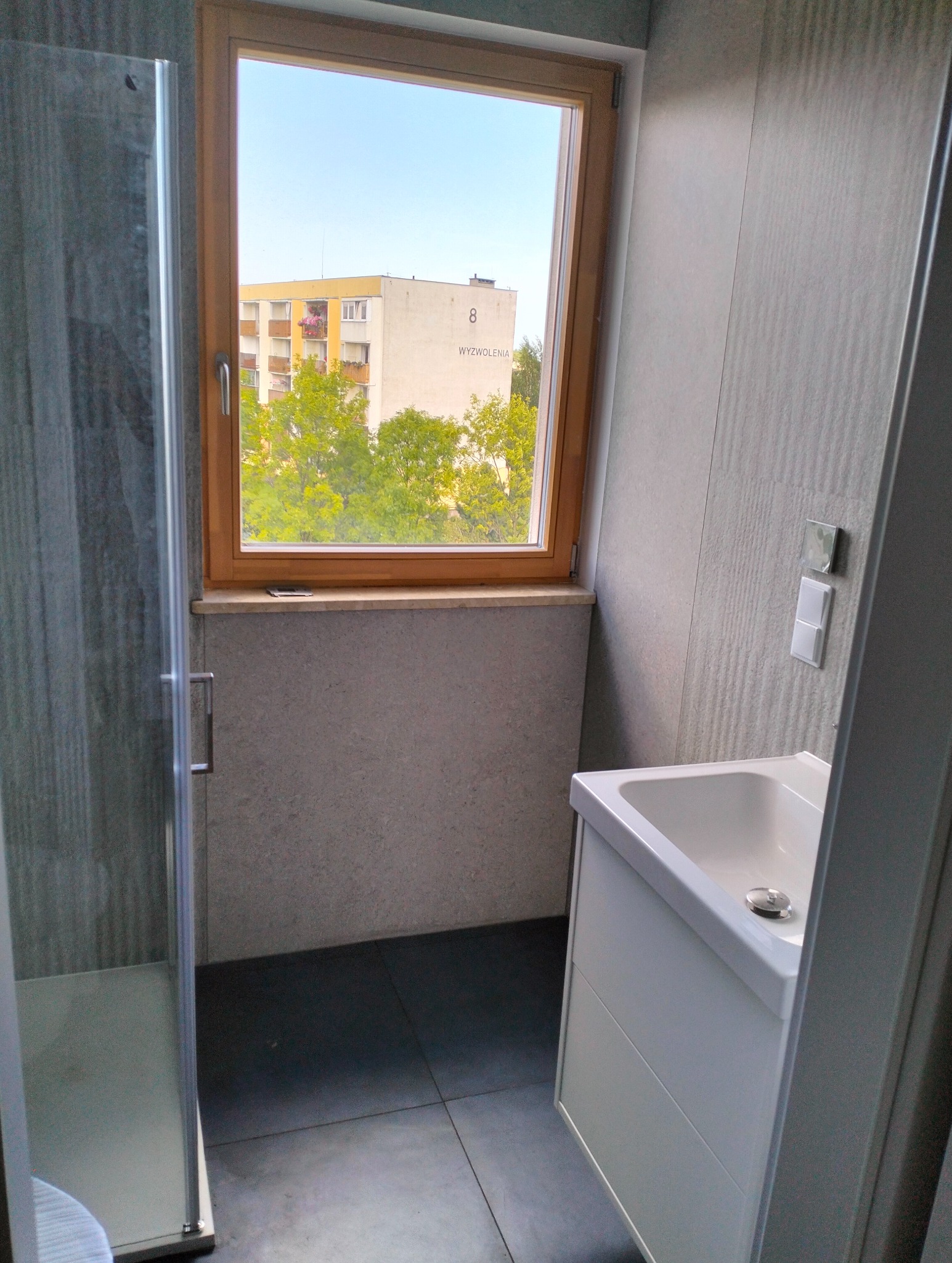 Wnętrze łazienki z kabiną prysznicową z szarego szkła, białą szafką z umywalką i drewnianym oknem z widokiem na blok mieszkalny.