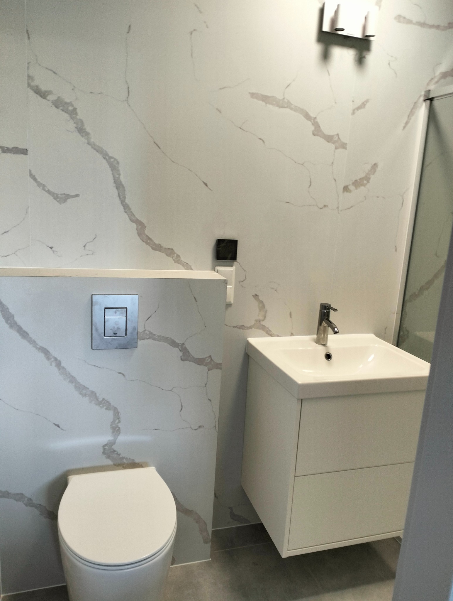 Nowoczesna łazienka z białą armaturą i płytkami imitującymi marmur, widoczna toaleta z chromowanym przyciskiem spłukiwania, umywalka z szafką i minimalistyczną baterią oraz fragment kabiny...