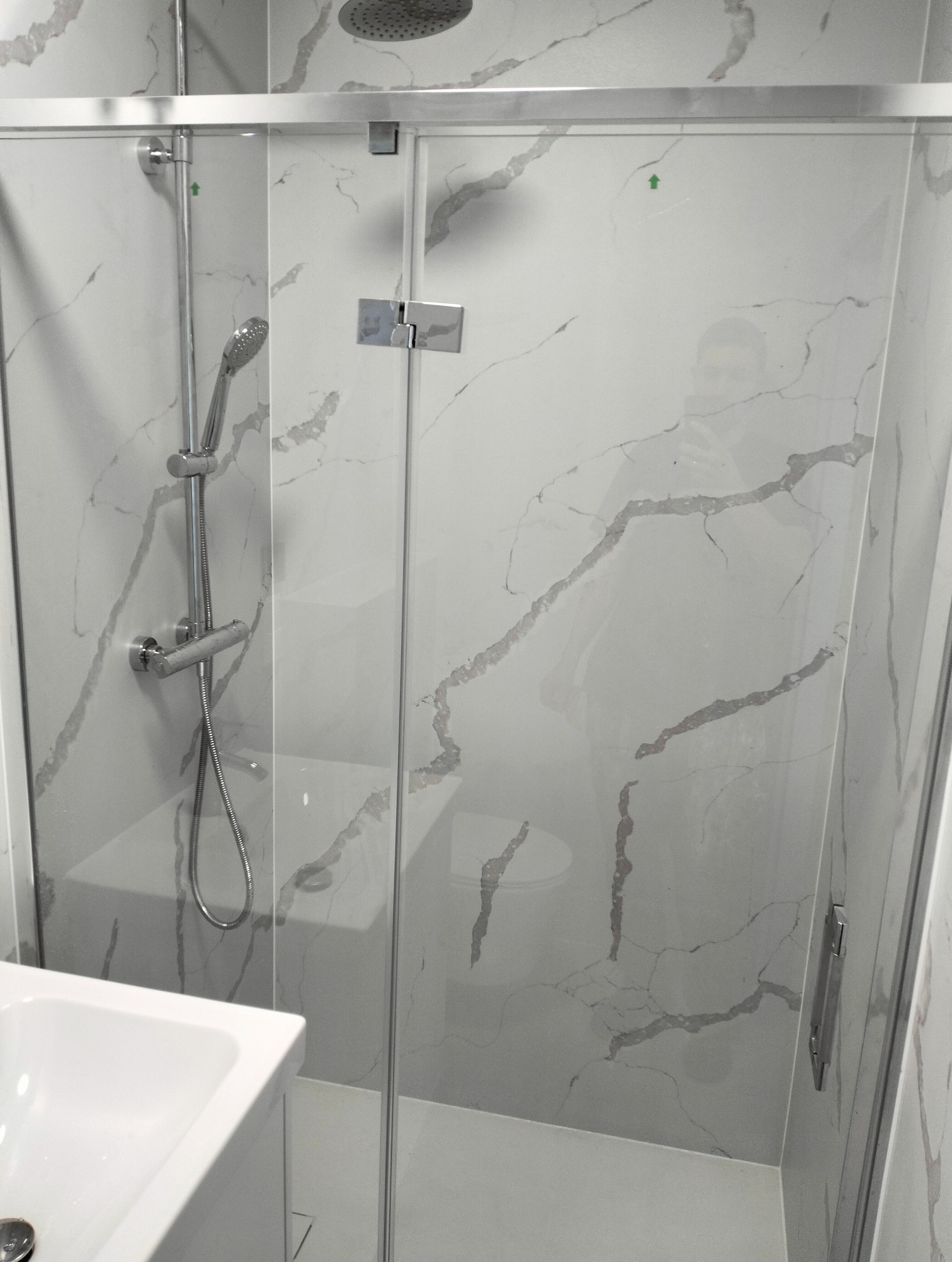 Nowoczesna kabina prysznicowa z deszczownicą i panelem imitującym marmur, widoczna umywalka po lewej stronie.