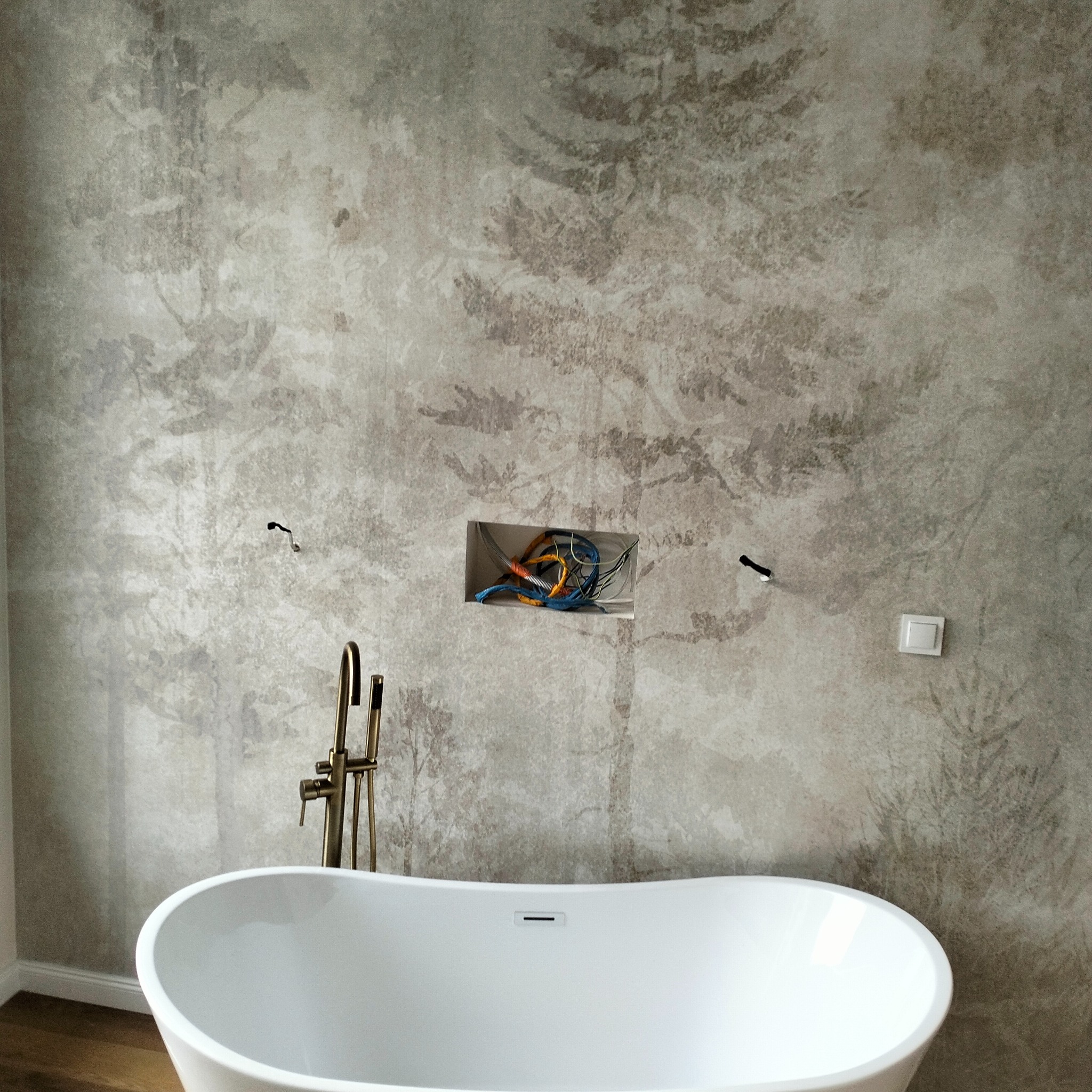 Biała wanna wolnostojąca z mosiężną armaturą na tle ściany z wzorem leśnym; widoczne przewody elektryczne w ścianie przed montażem osprzętu.