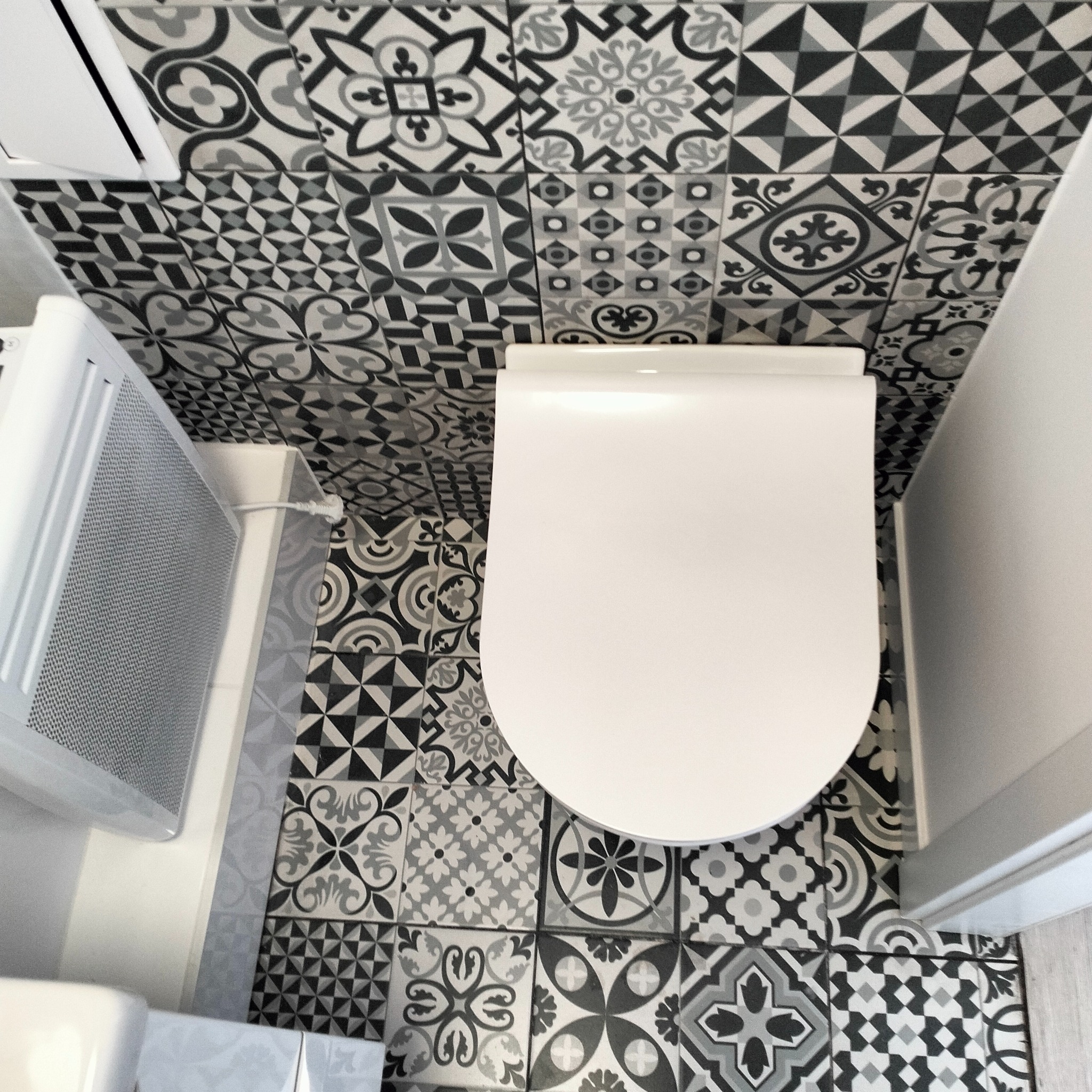 Toaleta z widokiem z góry na białą muszlę klozetową i grzejnik na tle ściany oraz podłogi wyłożonej czarno-białymi płytkami w różne wzory.