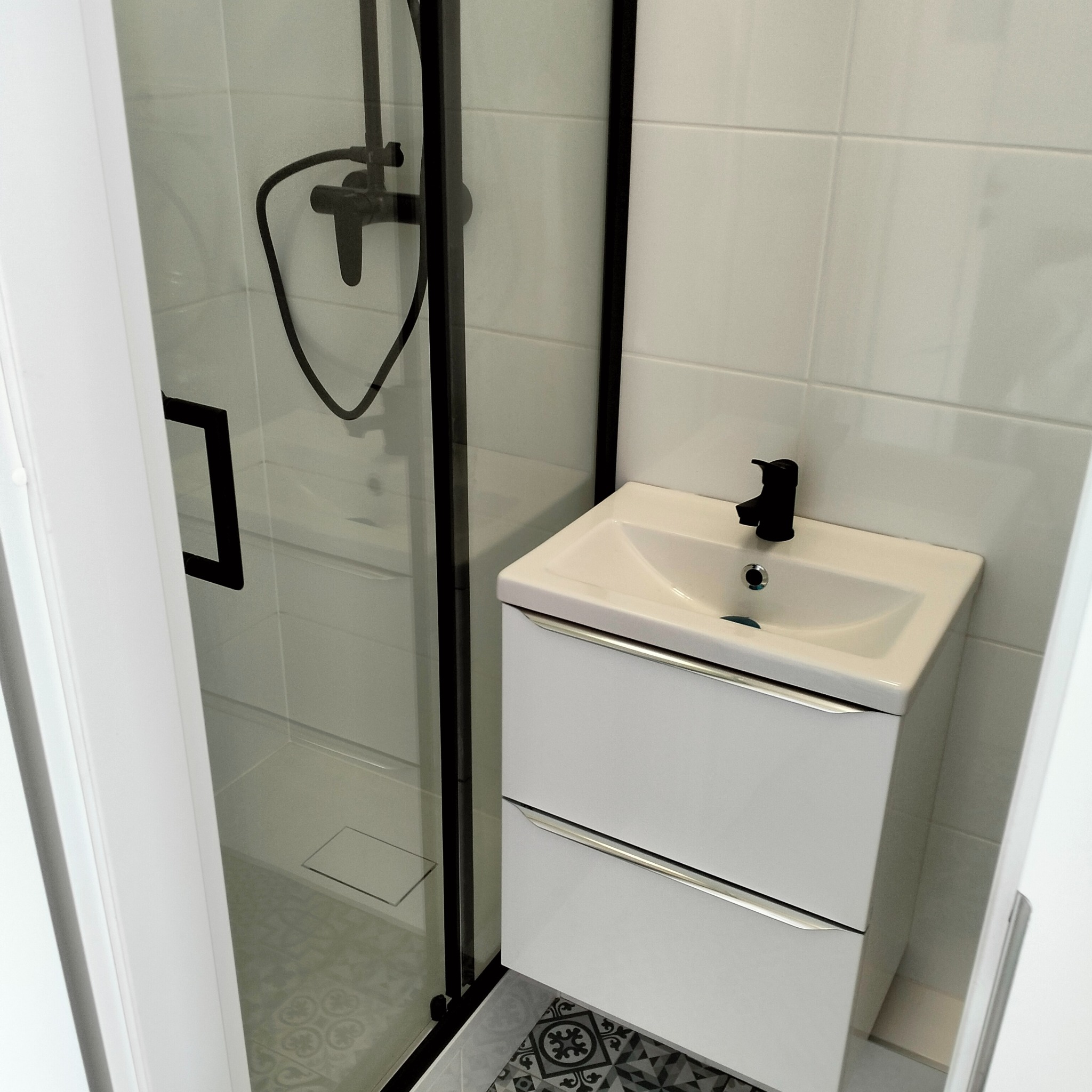 Nowoczesna łazienka z białą umywalką z szafką, czarną armaturą i kabiną prysznicową z czarnymi ramami, widoczne płytki podłogowe z geometrycznym wzorem.