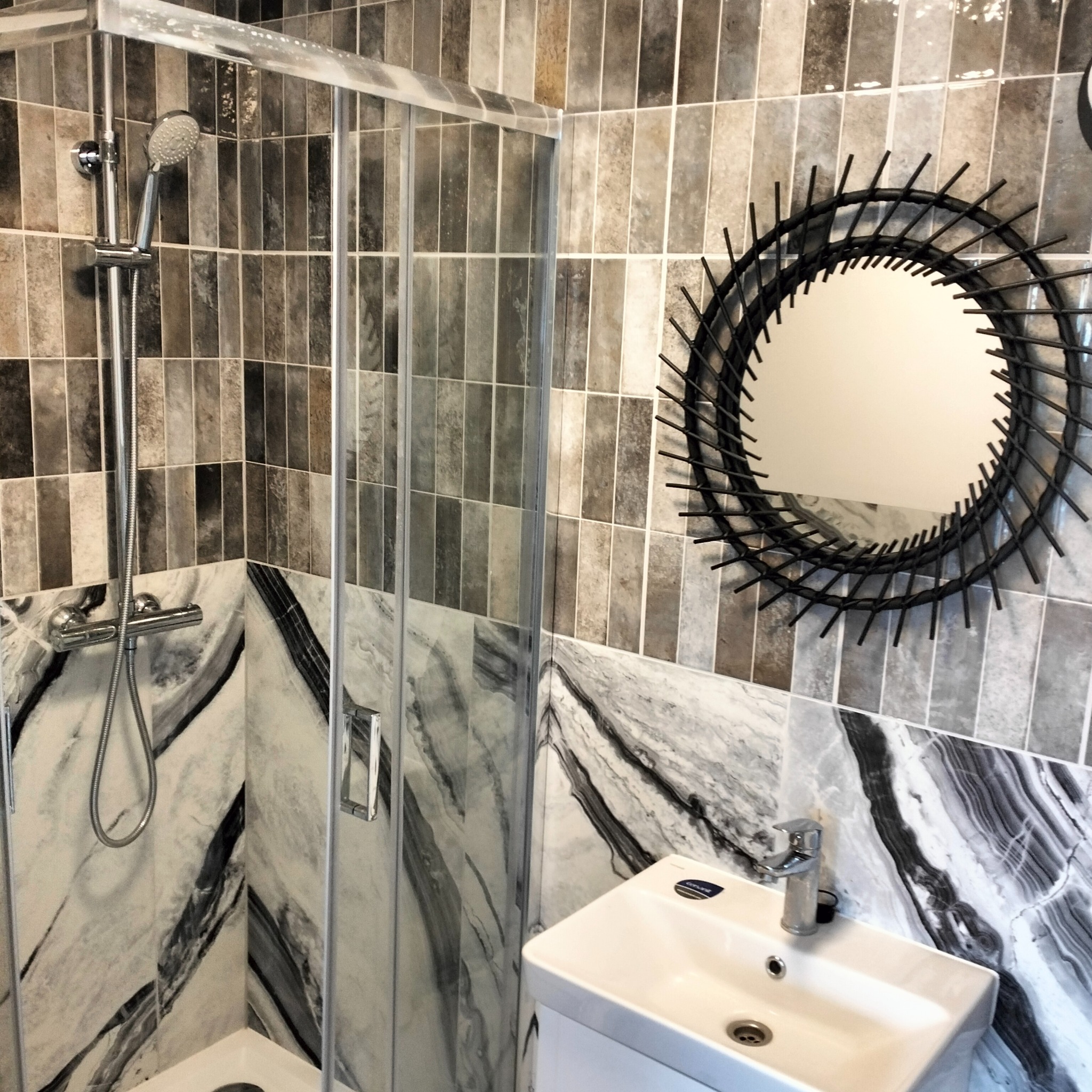 Nowoczesna łazienka z kabiną prysznicową ze szklanymi drzwiami, umywalką nablatową i okrągłym lustrem z czarną ramą imitującą słońce, wykończona płytkami w odcieniach szarości i bieli z marmurkowym...