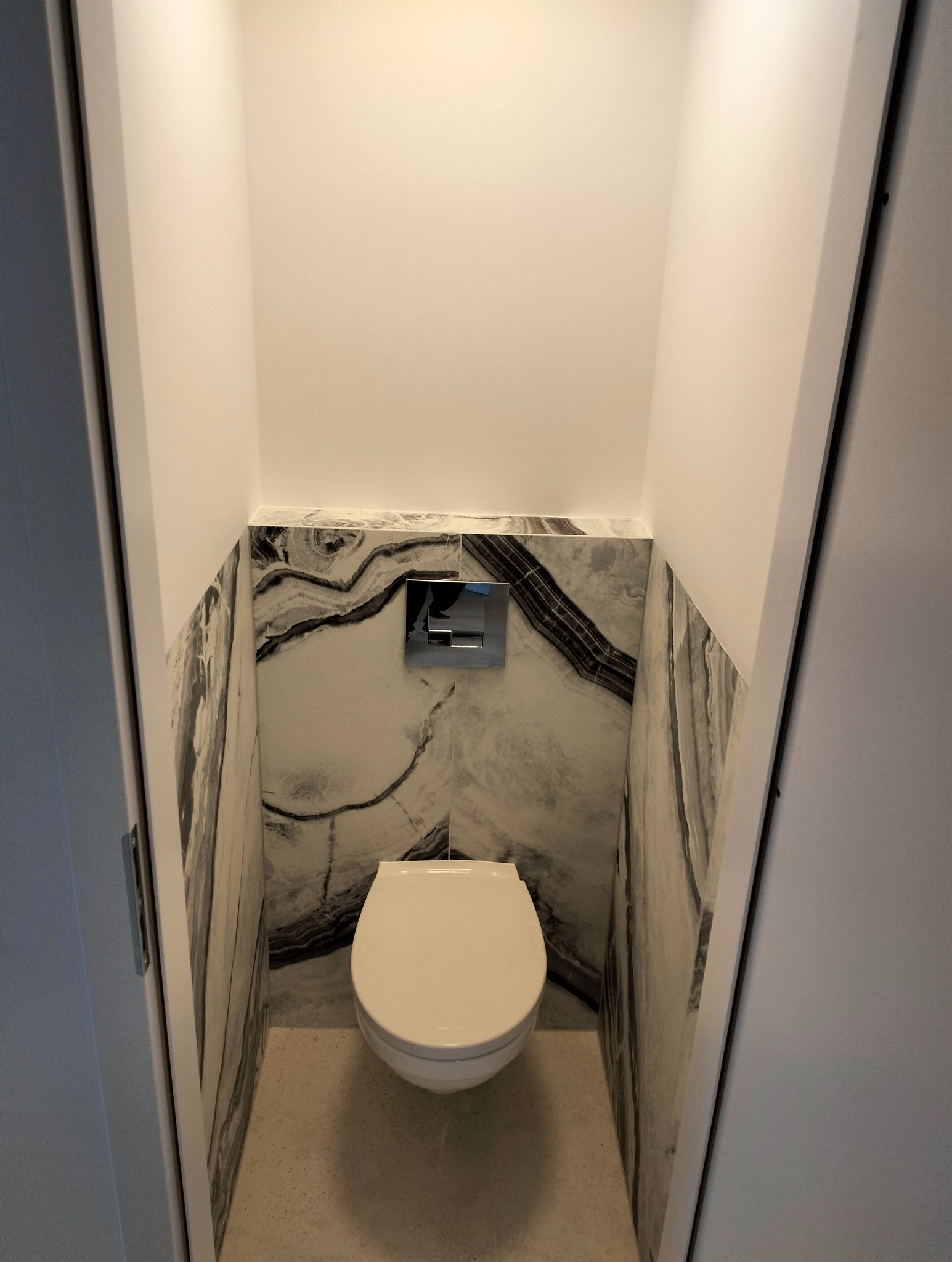 Kompaktowa toaleta w małej łazience z marmurowymi płytkami na ścianach i podwieszanym systemem spłukiwania.