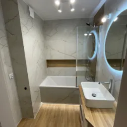 Nowoczesna łazienka z wanną za szklanym parawanem, umywalką nablatową i okrągłym lustrem z podświetleniem LED. Płytki imitujące marmur i drewnopodobna podłoga.
